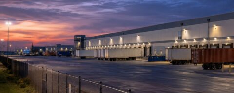 Warehouse security at a 3PL distribution center with loading docks, trailers, perimeter fencing and outdoor surveillance cameras at dusk.